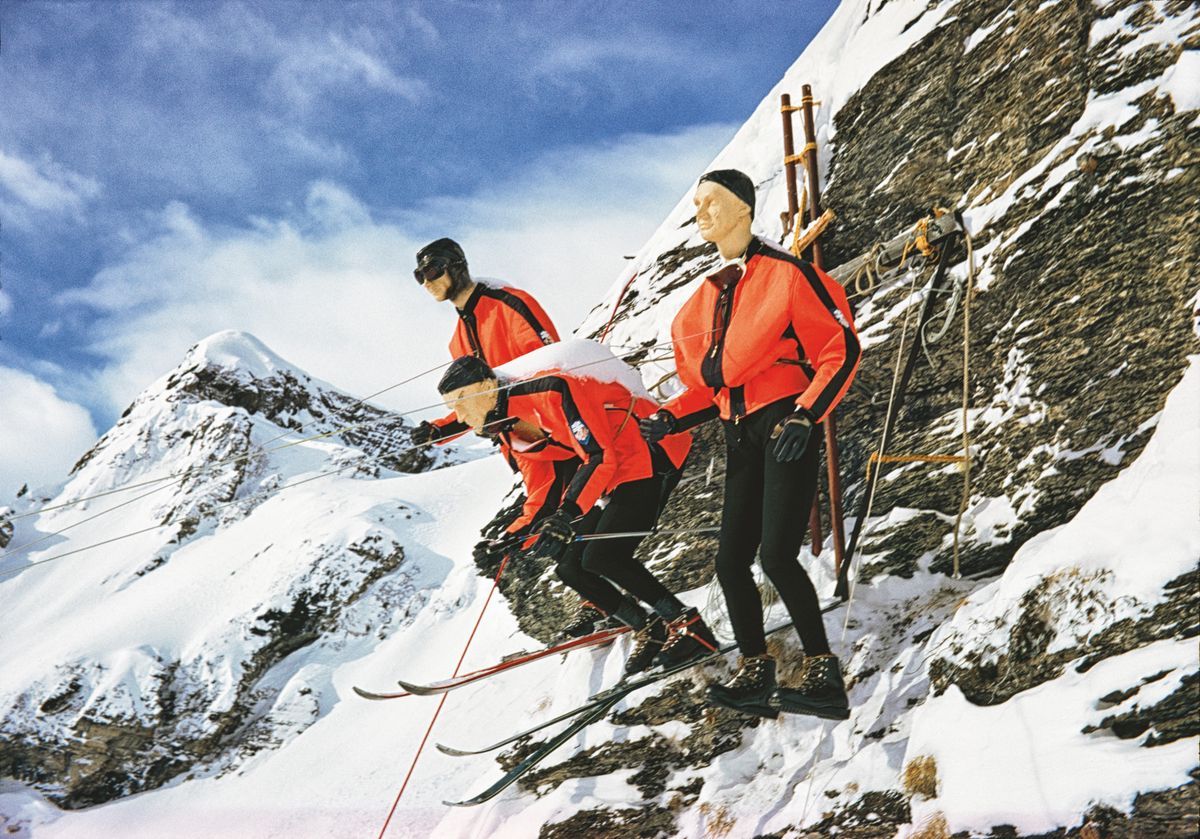 Three skiers in red combinations about to descend a snowy slope, with mountains in the background. Three skiers in red combinations about to descend a snowy slope, with mountains in the background.