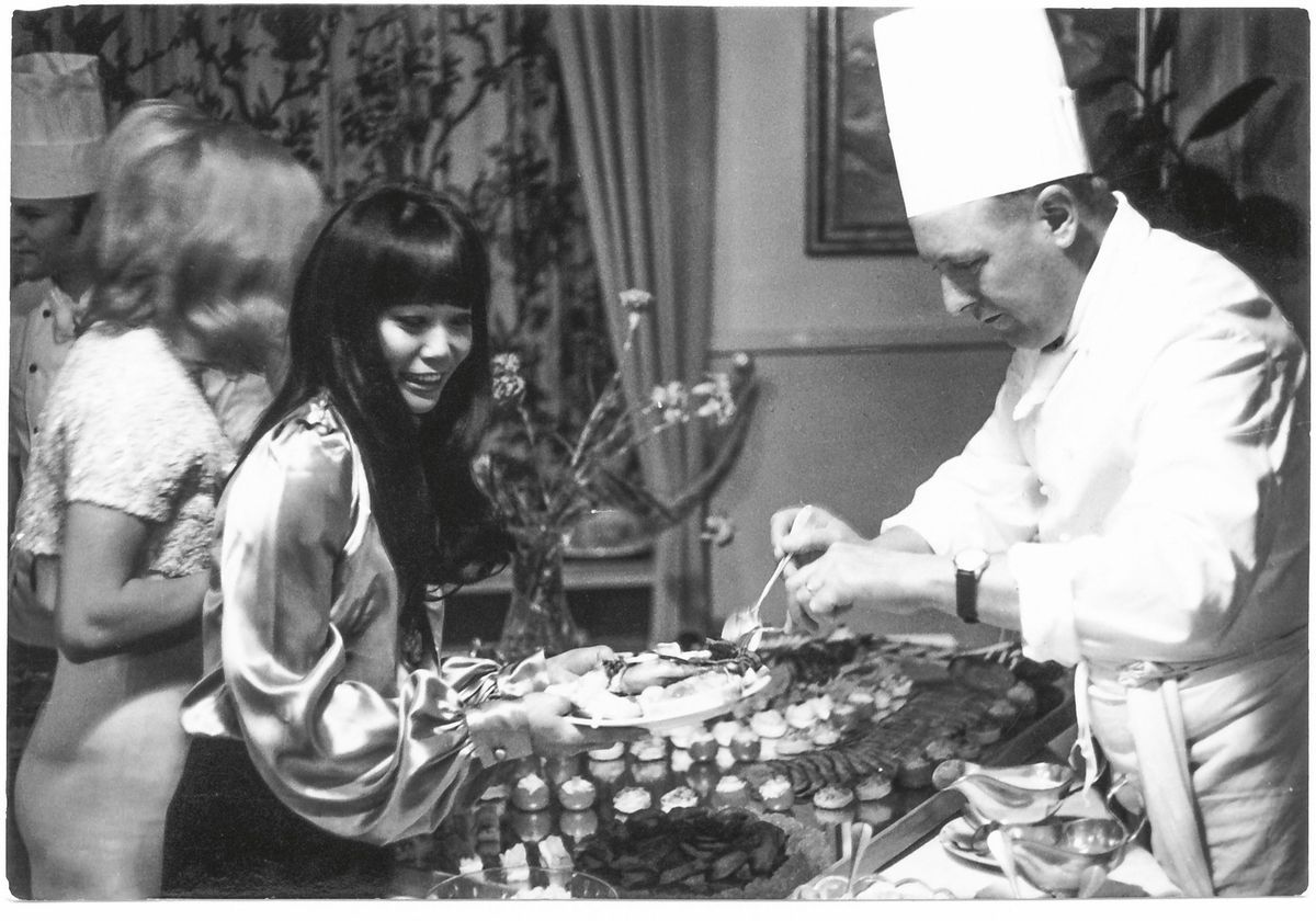 A chief in white clothes serves food for a smiling woman during an elegant buffet. A chief in white clothes serves food for a smiling woman during an elegant buffet.