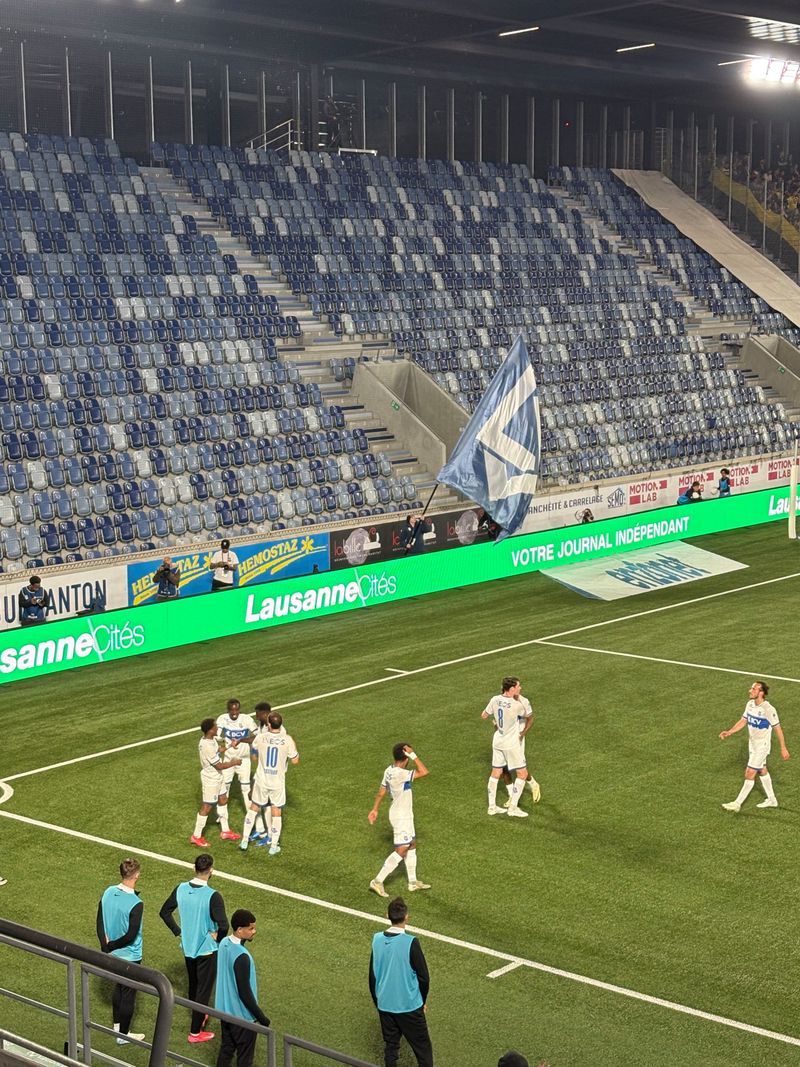 White football players celebrate a goal on the field, surrounded by replacements and a largely empty stadium. White football players celebrate a goal on the field, surrounded by replacements and a largely empty stadium.