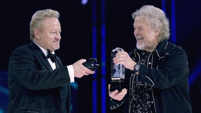 A man holds a small camera in front of the trophy exposed by another man on a stage, during a gala evening.