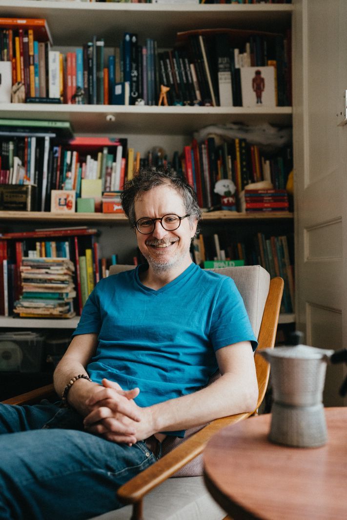 A smiling man wearing a blue t-shirt seated on a chair in front of a well-stocked library. A smiling man wearing a blue t-shirt seated on a chair in front of a well-stocked library.