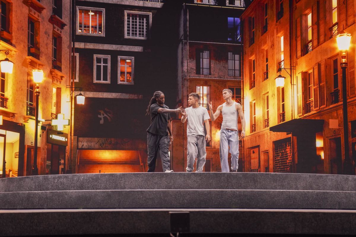 Three people walking on a theater scene representing an urban street at night, lit by lampposts. Three people walking on a theater scene representing an urban street at night, lit by lampposts.