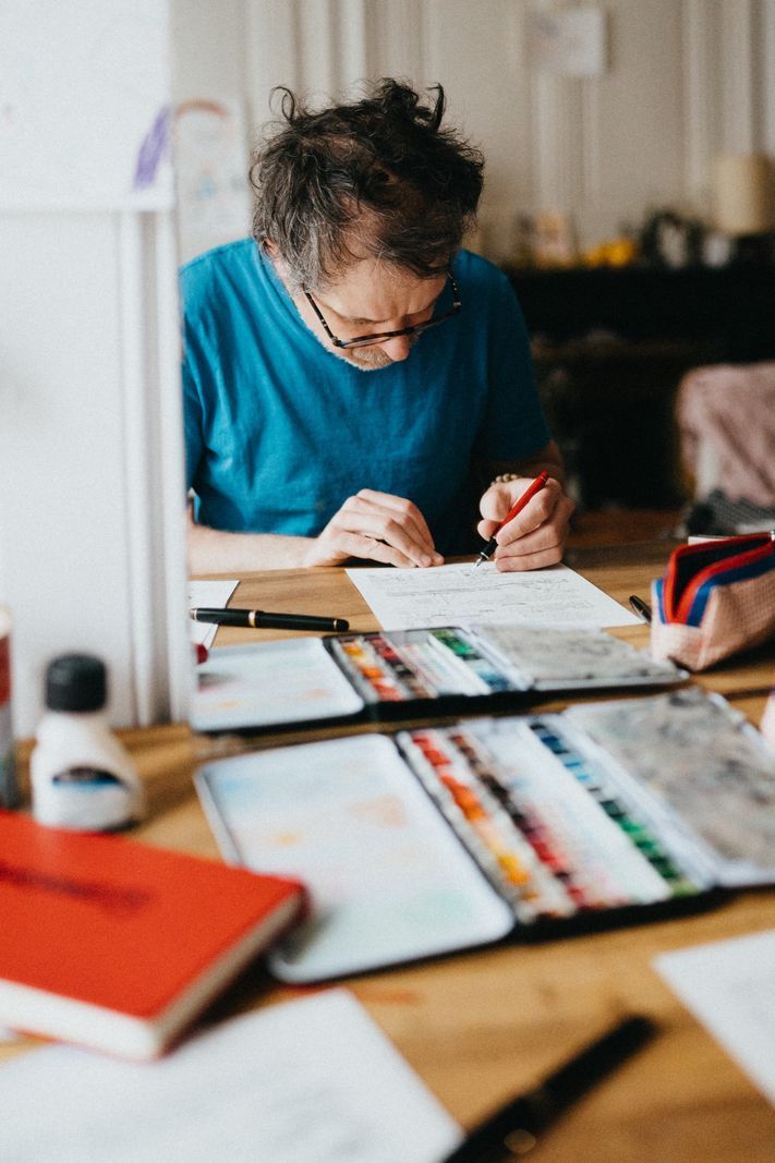 Concentrated man, dressed in a blue t-shirt, drawing on a desk cluttered with watercolors and artistic supplies. Concentrated man, dressed in a blue t-shirt, drawing on a desk cluttered with watercolors and artistic supplies.