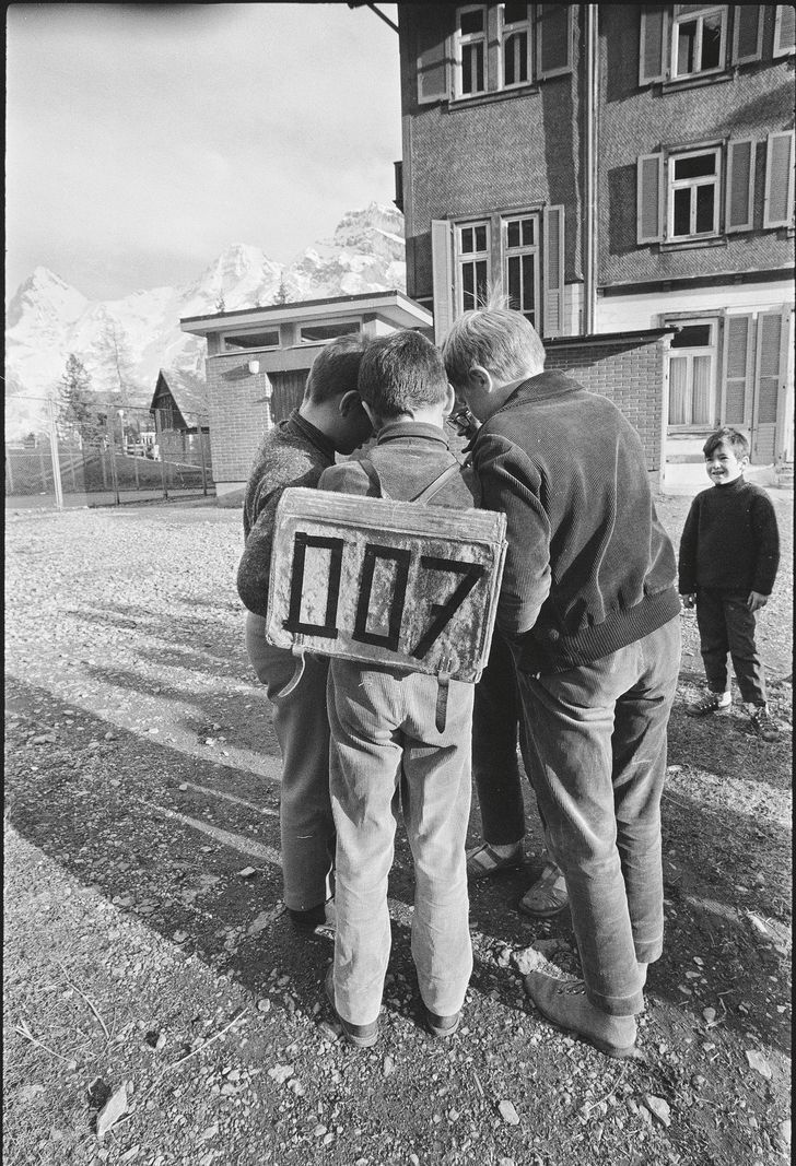 Children grouped around a marked panel '007' in a sunny courtyard, with mountains in the background. Children grouped around a marked panel '007' in a sunny courtyard, with mountains in the background.