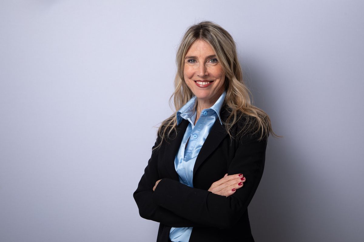 A smiling woman with blond and long hair, wearing a blue shirt and a black jacket, arms crossed in front of a gray background. A smiling woman with blond and long hair, wearing a blue shirt and a black jacket, arms crossed in front of a gray background.
