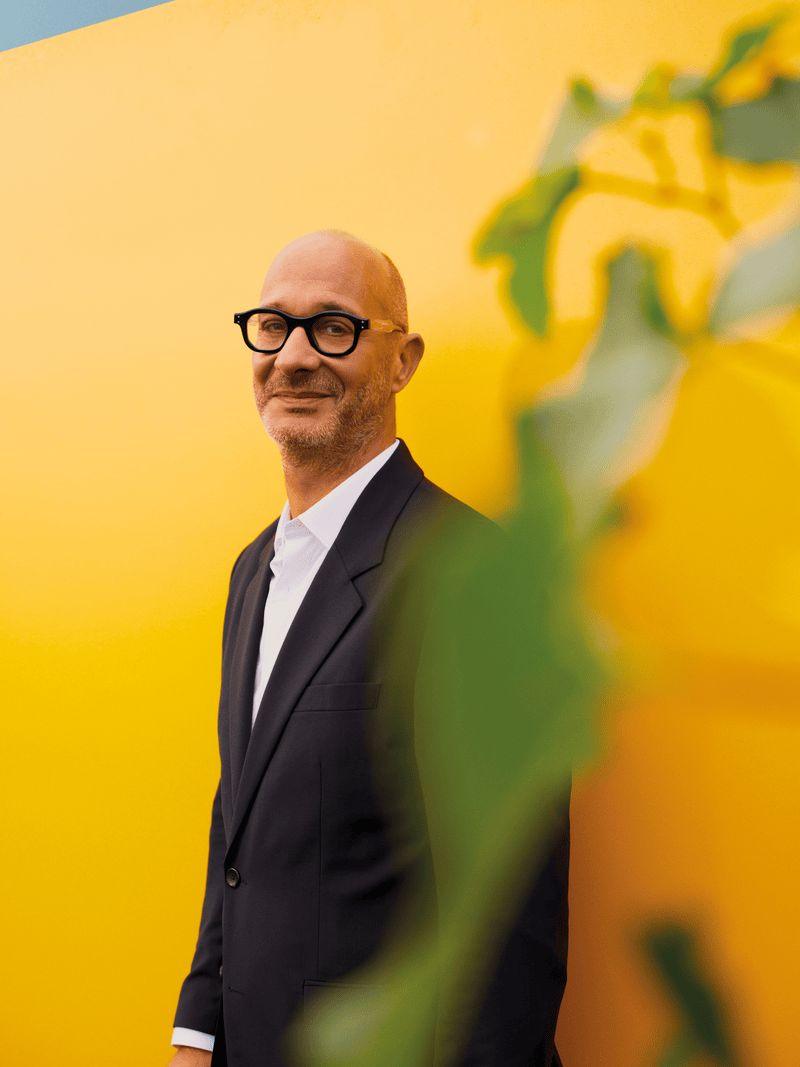 Man with black glasses, dressed in a dark suit, standing in front of a bright yellow wall with a blurred plant in the foreground. Man with black glasses, dressed in a dark suit, standing in front of a bright yellow wall with a blurred plant in the foreground.