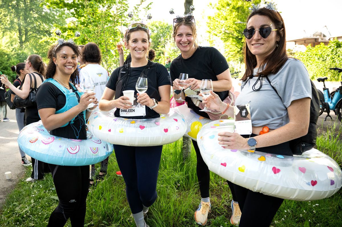 Four participants in the Apero Run de Lausanne carrying buads and holding glasses of wine, smiling in a green setting. Four participants in the Apero Run de Lausanne carrying buads and holding glasses of wine, smiling in a green setting.