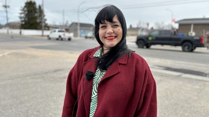 A woman wearing a microphone to her coat smiles on the camera outside.