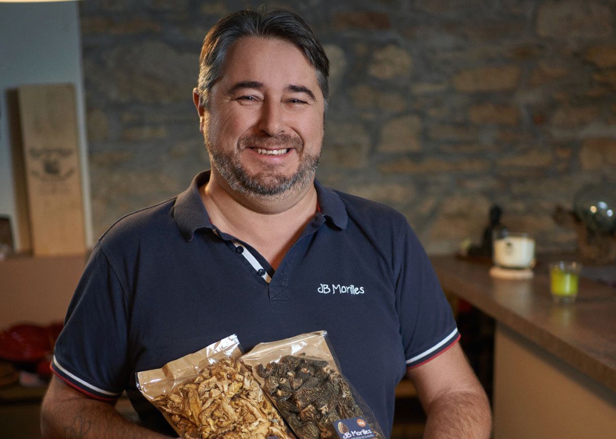 A smiling man holds two sachets of food products in front of a stone wall, in a warm interior. A smiling man holds two sachets of food products in front of a stone wall, in a warm interior.