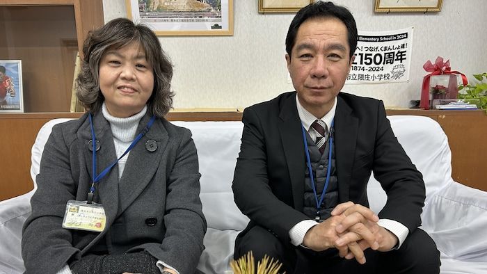 A man and a woman sit on a couch and watch the camera.