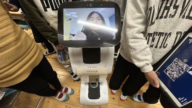 A robot whose head is a screen that displays a young girl's face is surrounded by students.